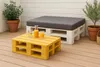 A patio setting with a yellow pallet coffee table and a white pallet bench with a gray cushion. There are two glasses of orange juice and a bottle of orange juice on the coffee table. Two potted plants are also visible. Anteprima dell’immagine prodotto