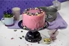 A pink cake decorated with blueberries and pansies sits on a cake stand, surrounded by baking ingredients and utensils. Produktbild-Vorschau