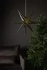 A dark green, seven-pointed star lantern hangs in front of a dark gray wall, next to a Christmas tree and a dark brown sofa. Podgląd zdjęcia produktu