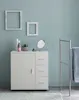 A white bathroom cabinet with a door and three drawers, a mirror, and some toiletries on top, stands against a light blue wall. A towel rack with a blue towel is to the right. Náhled obrázku produktu