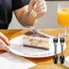 A slice of tiramisu cake on a white plate, with a person holding a gold fork above it and two black sporks with faces on the plate. A glass of orange juice is in the background. Produktbild-Vorschau
