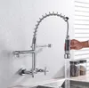 A chrome kitchen faucet with a pull-down sprayer is shown mounted on a tiled wall above a sink. A hand is holding the sprayer, and water is running into the sink. Two jars of coffee beans are visible in the background. Podgląd zdjęcia produktu