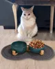 A cat sits under a table, looking at a set of pet bowls filled with water and food, placed on a mat. Produktbild-Vorschau