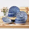 A collection of blue and white porcelain tableware is arranged on a wooden surface, with a vase of pink flowers and gift boxes in the background. Produktbild-Vorschau