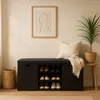 A black shoe cabinet with a padded seat, featuring shelves for shoes, a woven basket, a framed picture on the wall, and a potted plant on a stool. Náhľad obrázku produktu