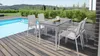A patio dining set next to a swimming pool on a wooden deck, with a landscaped background. Produktbild-Vorschau