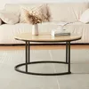 A round wooden coffee table with black metal legs is placed on a rug in front of a sofa. A vase with dried flowers and a stack of books are on the table. Produktbild-Vorschau