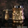 A close-up studio shot of a decorative mug with a metallic gold finish, featuring intricate detailing and a handle shaped like a gauntlet. The mug is placed on a reflective surface against a dark, textured background. Podgląd zdjęcia produktu