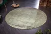 A round, distressed green rug is placed on a dark wooden floor in a room setting. A green sofa leg and a decorative tray with a cup are visible in the background. Produktbild-Vorschau