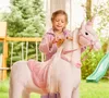 A young girl is brushing the mane of a large, plush unicorn toy in a backyard setting. Produktbild-Vorschau