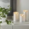Three LED candles of varying heights are displayed on a white surface, with a potted plant to the left and a framed picture in the background. Podgląd zdjęcia produktu