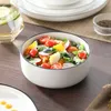 A close-up of a salad in a white bowl with a black rim, set on a wooden table. Other tableware and a glass of water are visible in the background. Náhled obrázku produktu