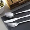 A set of cutlery, including a spoon, knife, and fork, is laid out on a dark slate surface. In the background, a white bowl with quail eggs and a wooden tray are visible. Náhled obrázku produktu
