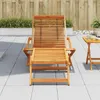 A wooden lounge chair with a footrest is set up on a tiled patio, with a small wooden side table to the right. A wooden fence and some greenery form the background. Náhľad obrázku produktu