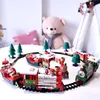 A festive toy train set with Christmas-themed decorations, including Santa Claus figures and miniature trees, arranged on a track. A large teddy bear and a teepee are visible in the background. Náhľad obrázku produktu