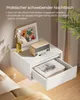 A white floating nightstand with a drawer open, displaying books and glasses, next to a bed. A small wooden radio and a vase with flowers are on top of the nightstand. Produktbild-Vorschau