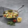 A stainless steel frying pan filled with pasta, tomatoes, and spinach, placed on a gray surface next to a folded gray napkin, a tomato, spinach leaves, and a nest of uncooked pasta. Podgląd zdjęcia produktu