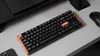 A black mechanical keyboard with an orange accent key and a wooden-like bezel is placed on a light gray surface, with a black mouse to the right and a corner of a smartphone to the left. Podgląd zdjęcia produktu