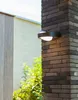 An outdoor wall light mounted on a brick pillar, with greenery and a building in the background. Produktbild-Vorschau