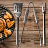 A set of stainless steel barbecue tools, including a spatula, tongs, and fork, are laid out on a wooden surface next to a grill with food. Anteprima dell’immagine prodotto
