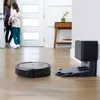 A robot vacuum cleaner and its charging dock are on a wooden floor in a hallway, with two people entering through a doorway in the background. Voir les images du produit