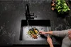 A person is rinsing vegetables in a kitchen sink with a black faucet and a black sink. Produktbild-Vorschau