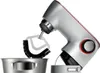 A close-up shot of a silver stand mixer with a red accent ring around the dial, positioned over a stainless steel bowl. The mixer's attachment, a black and white beater, is visible. Anteprima dell’immagine prodotto