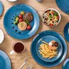 A table setting with blue ceramic plates, bowls, and cups, featuring food items like steak, pasta, salad, and french fries. Náhled obrázku produktu