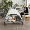 A woman sits on a chair on a patio next to a raised dog bed with a canopy, where two dogs are resting. Náhľad obrázku produktu