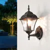 A vintage-style outdoor wall lamp mounted on a white wall, with a garden scene in the background. Produktbild-Vorschau