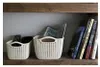 Two cream-colored woven storage baskets are on a wooden shelf. One basket contains a remote control and a black object. The other basket contains a magazine. Several books are standing upright next to the baskets. Náhľad obrázku produktu