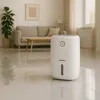 A white dehumidifier is placed on a tiled floor in a living room with a sofa and a plant in the background. Podgląd zdjęcia produktu