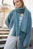 A woman wearing a blue knitted cardigan and scarf stands on stone steps. Produktbild-Vorschau