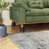 Close-up of a green corduroy sofa with black furniture risers under its legs, placed on a wooden floor next to a rug and a potted plant. Produktbild-Vorschau