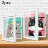 Two open white frames displaying cartoon animal artwork, placed on a wooden surface against a white brick wall. Produktbild-Vorschau