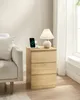 A bedside table with a lamp, phone, and earbuds on top, next to a couch. Náhled obrázku produktu