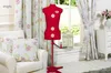 A sewing mannequin is placed next to a chair with cushions and curtains in a room with a window. Produktbild-Vorschau
