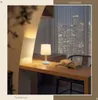 A bedside lamp is placed on a wooden desk next to an open book, with a city skyline visible through the window in the background. Náhľad obrázku produktu