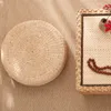 A round straw cushion and a rectangular woven tray with prayer beads and a small vase with red flowers are placed on a textured tan surface. Produktbild-Vorschau