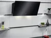 A black angled kitchen hood with integrated lighting, mounted on a white tiled backsplash above shelves with kitchenware and decorative plants. Voir les images du produit
