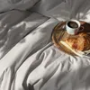 A breakfast tray with coffee and a croissant is placed on a rumpled grey bed. Produktbild-Vorschau