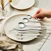 A hand is placing a small spoon on a plate with other cutlery. Voir les images du produit