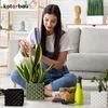 A woman is watering a snake plant in a decorative pot, surrounded by other plant pots and gardening tools. Produktbild-Vorschau