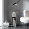 A narrow gray nightstand with a wooden top sits next to a stack of books and a candle, with a modern lamp overhead and a fluffy white chair to the side. Produktbild-Vorschau