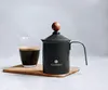 A black milk frother with a wooden knob on top sits next to a glass of coffee on a wooden board. Produktbild-Vorschau