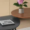 Two round coffee tables, one black and one brown, with a book on the black table and a potted pink flower on the brown table, against a light wood paneled background. Produktbild-Vorschau