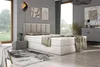 A bedroom scene featuring a beige boxspring bed with a headboard, bedside table, rug, and artwork on the wall. Produktbild-Vorschau