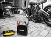 A motorcycle battery charger and battery are shown in a workshop setting with a motorcycle in the background. Podgląd zdjęcia produktu