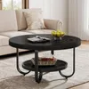 A round, black coffee table with a lower shelf is placed in a living room setting, next to a beige sofa. The table is decorated with a book, a bowl of fruit, and a basket with decorative items. Produktbild-Vorschau