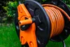 A close-up of an orange garden hose wound around a black and orange hose reel, set against a blurred green background of grass and foliage. Voir les images du produit